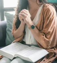 A woman with a Bible in her lap with clasped hands praying.