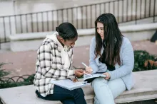 Two young women sitting outside on a bench and writing in notebooks