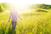 A woman walking in field of tall grass and flowers as sun rays shine on her.