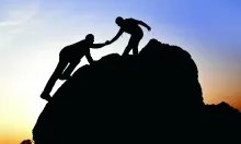 Silhouette of a person helping another person climb a boulder.