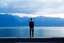 a man wearing black standing with his back to the viewer as he faces a blue mountainous landscape over a body of water