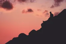 A person sitting a hill with a red sky.