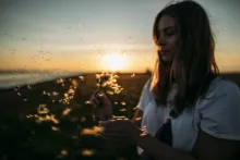 A woman holding a dandelion with the seeds of the plant blowing away.