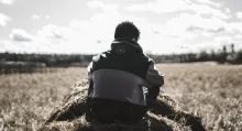 A young man sitting on a round bale of hale looking out over a field.