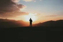 A silhouette of a person stand on a hill with the sun in the background.