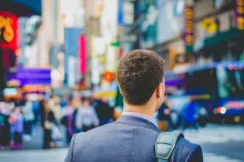 A young man wearing a suit and carrying a leather case on his shoulder walking in busy city.