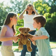 Two siblings fight over a stuffed teddy bear with the mom in the background.