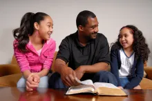 A dad and his two daughters talking with a Bible opened.