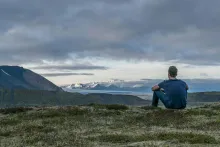 A young man sitting on the grass looking at a view of mountains.