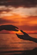 one hand reaching for another silhouetted against a sunset over water