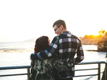 a man with his arm around a woman as they look at each other and stand by a railing overlooking a body of water