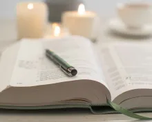 a bible with a pen lying across its open pages and candles and a teacup in the background