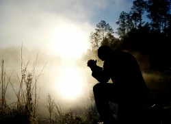 a silhouette of a praying man outdoors among shadowy trees and a morning mist