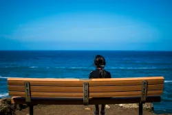 A woman sitting on a bench looking out on a large lake.