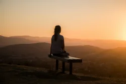A young woman sitting on a bench looking out over a vista of rolling hills.