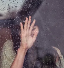 a sad woman with her hand up against a pane of glass dotted with raindrops