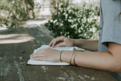 a woman's arms resting on an open Bible outdoors on a wooden table