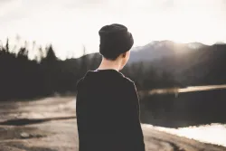 a boy facing away from the camera towards a landscape of water, sand and hills