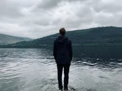 a man turned away to face a body of water with green mountains and clouds in the distance