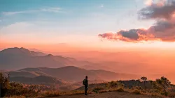 a man standing in a field looking towards a sunset landscape with mountains