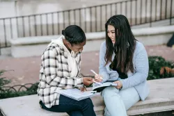 Two young women sitting outside on a bench and writing in notebooks