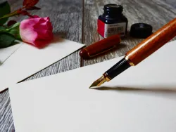 a fountain pen, bottle of ink, blank page, envelope and rose on a table