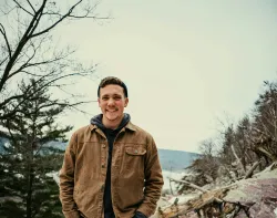 a smiling young man standing outdoors with a mountainous background