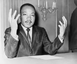 Rev. Martin Luther King, head-and-shoulders portrait, seated, facing front, hands extended upward, during a press conference.