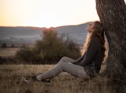 A young woman sitting on the ground leaning against a tree looking up toward the sky.