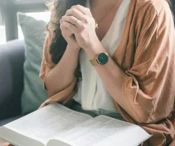 a woman sitting with hands folded in prayer with an open Bible in her lap