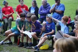 a group of people outdoors around a campfire, with a group of boys gathered around a song sheet