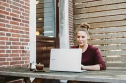 a woman seated at a wooden bench with a laptop and coffee with a brick wall and a door in the background