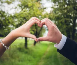 a woman's hand and a man's hand joined to make a heart shape with a green field and trees in the background