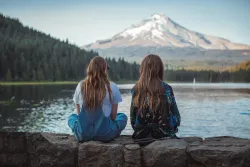 two women sitting on a ledge and looking at a lake