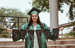 a young woman wearing a green graduation cap and gown while holding a banner that reads "senior."