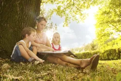 A woman sitting outside beneath a tree reading to her two children.