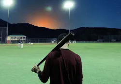 A young man holding a baseball bat on an empty ballfield at dusk.