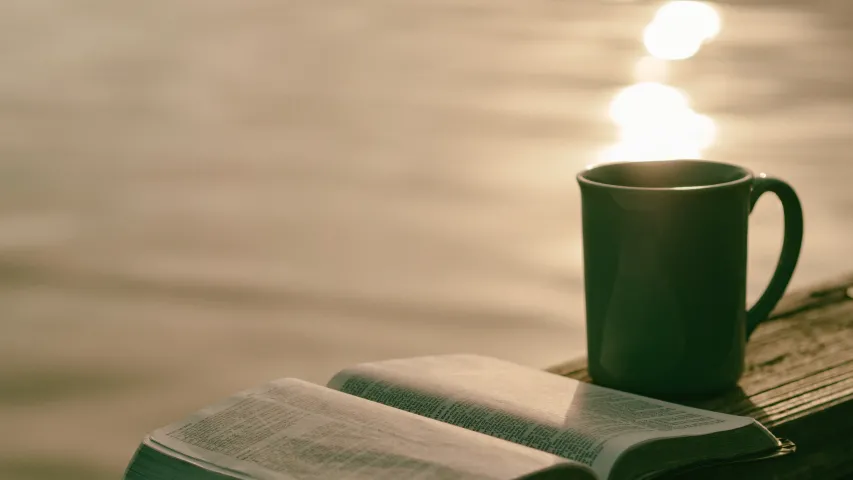An open Bible and a ceramic mug sitting on a table with a pond in the background.