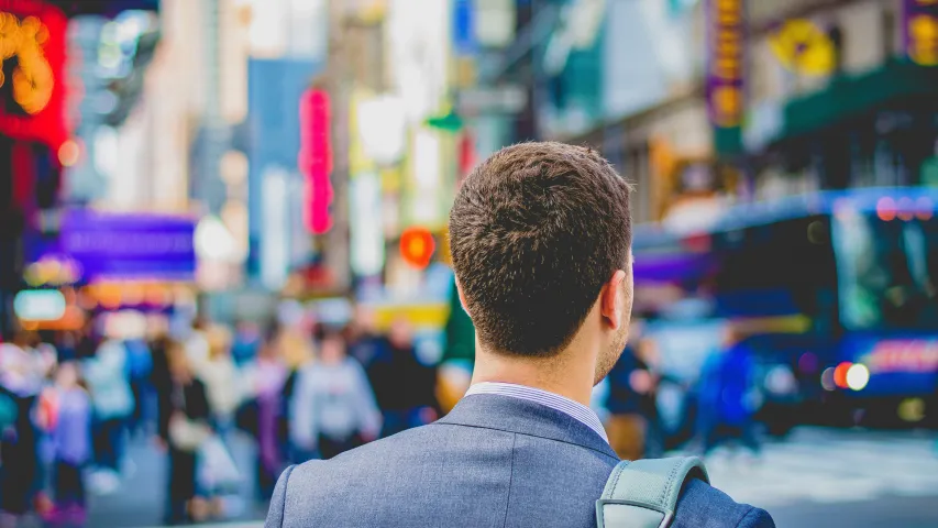 A young man wearing a suit and carrying a leather case on his shoulder walking in busy city.